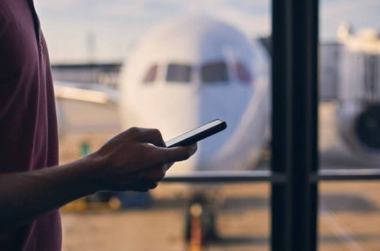 A person stands at the airport, holding a phone near a plane, contemplating, What is airplane mode used for before boarding.